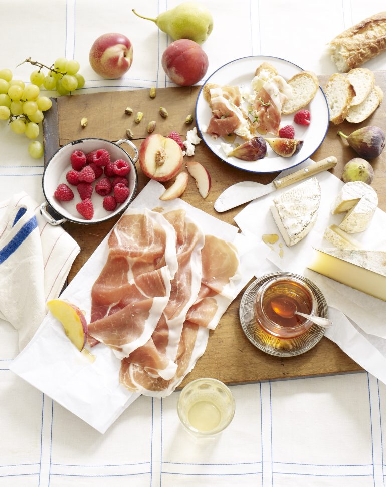 Charcuterie board on a picnic blanket with slices of Prosciutto di Parma, crusty bread, raspberries, grapes, pears, apples, honey and assorted cheeses